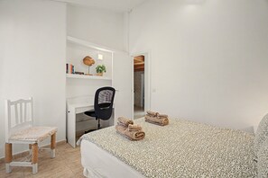A bedroom with a bed, a desk, a chair, and a small table with books and a plant.