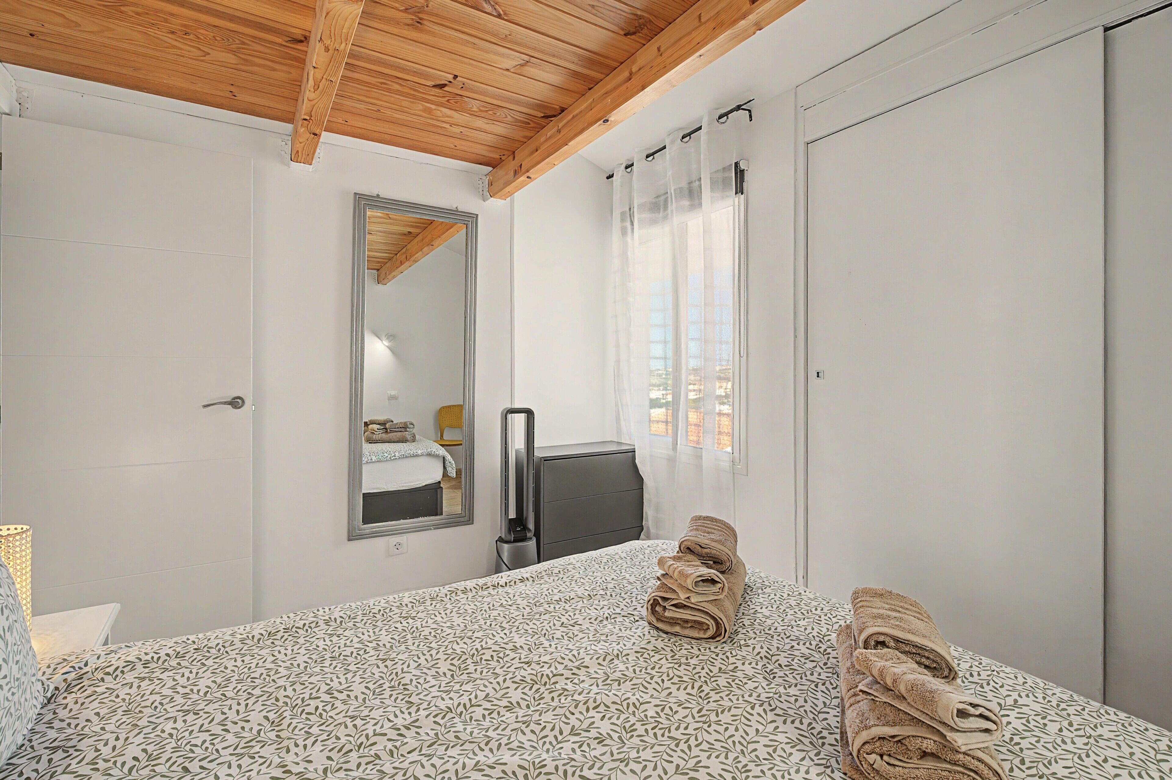 Un dormitorio con cama, espejo, ventana con cortinas y techo de madera.