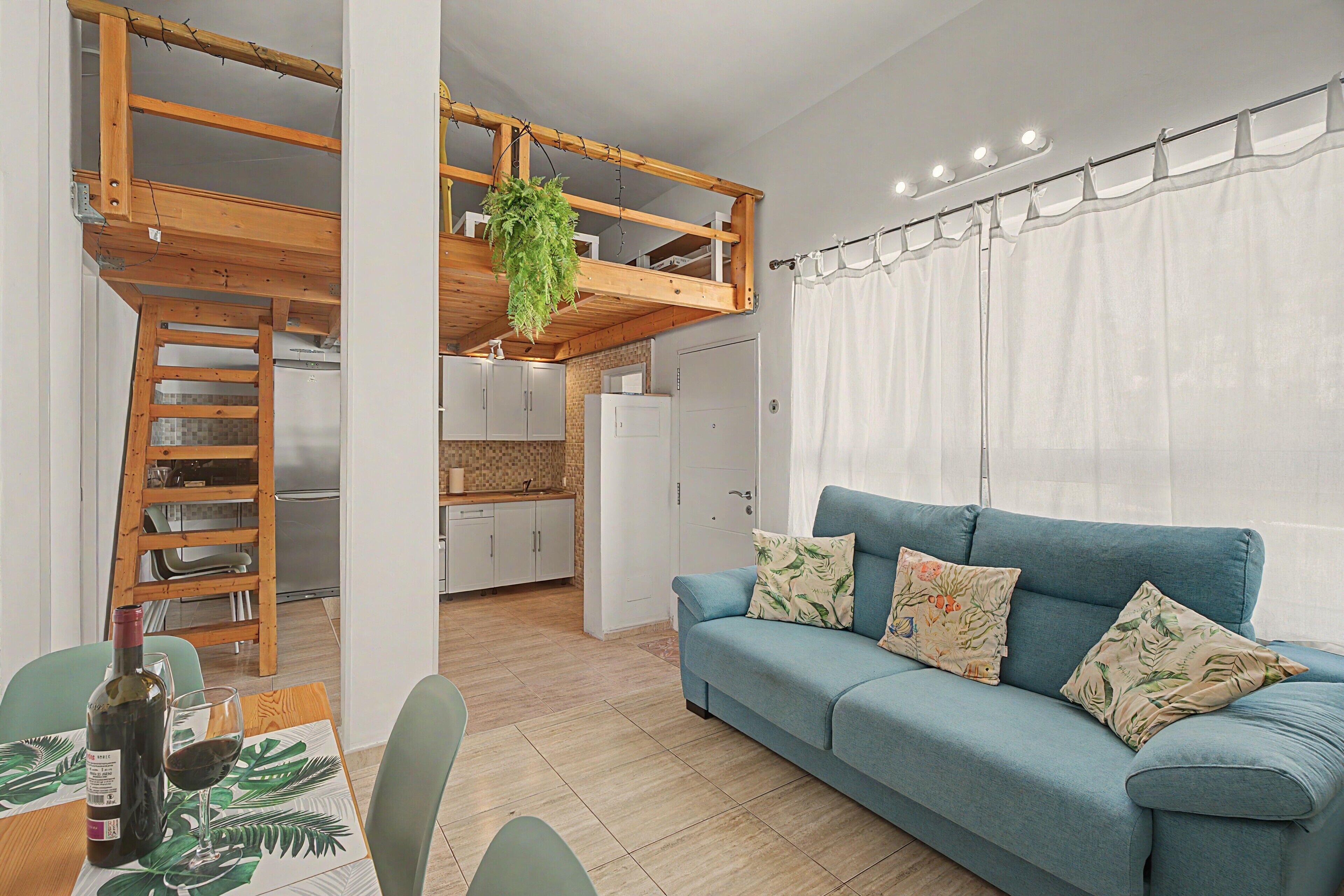 A modern living room with a blue sofa, a dining table with chairs, a kitchen area, and a loft space with a hanging plant.