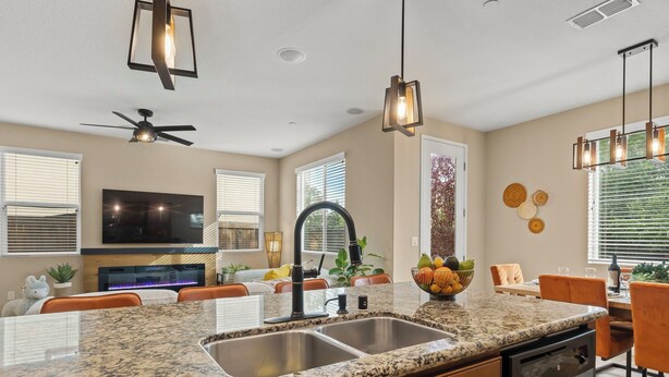 A modern kitchen with granite countertops, a double sink, and a dishwasher. There is a dining area with orange chairs and a large window.