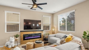 A living room with a flat-screen TV, a fireplace, a ceiling fan, and large windows with blinds.