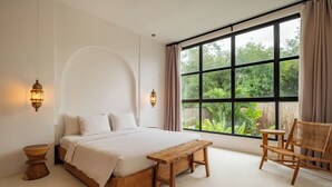 Un dormitorio con una cama grande, un banco de madera y una silla, una ventana con vistas a la vegetación y luces fijadas a la pared.