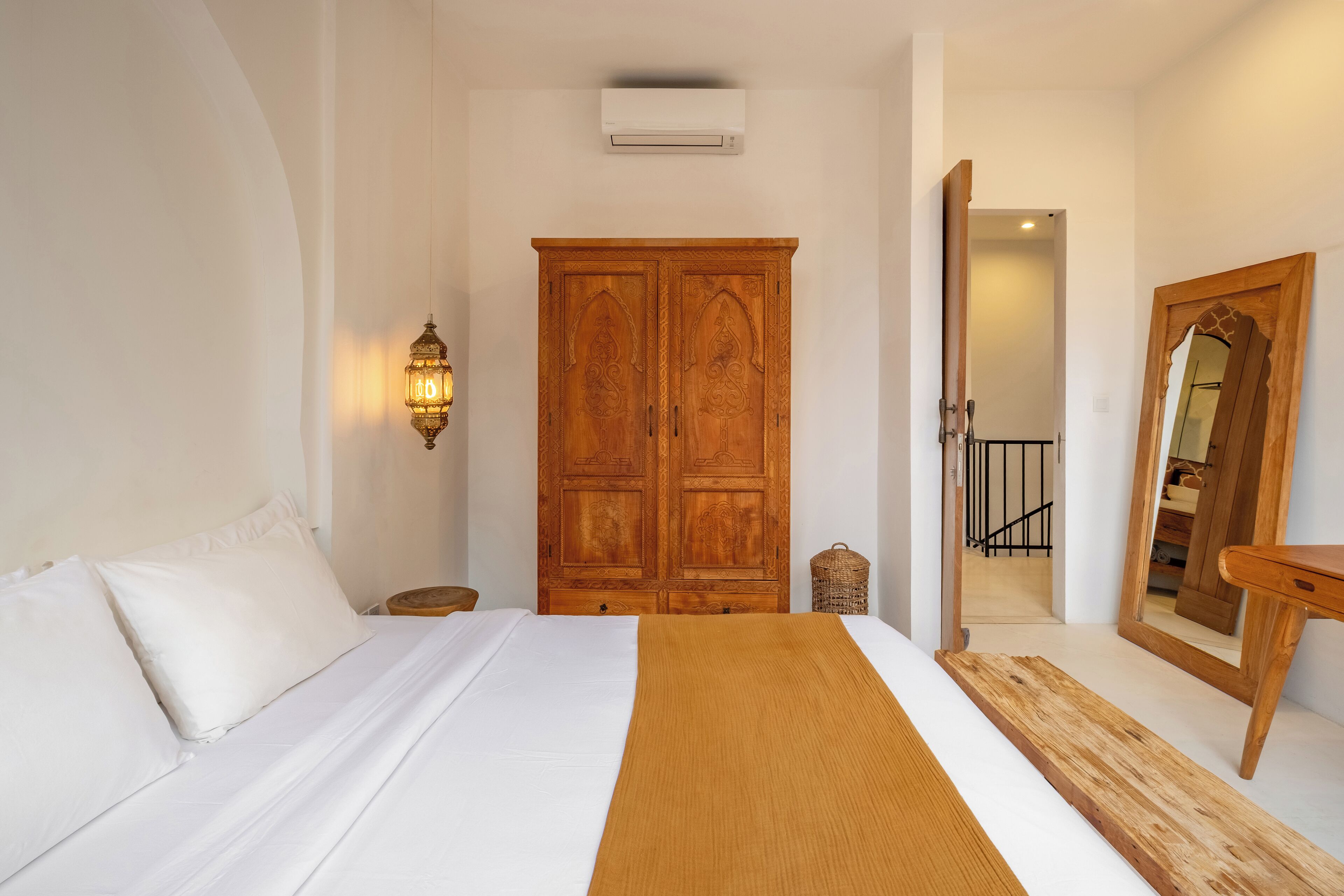A bedroom with a large wooden wardrobe, a bed with white bedding, a wooden bedside table, a wall-mounted light fixture, and a mirror with a wooden frame.