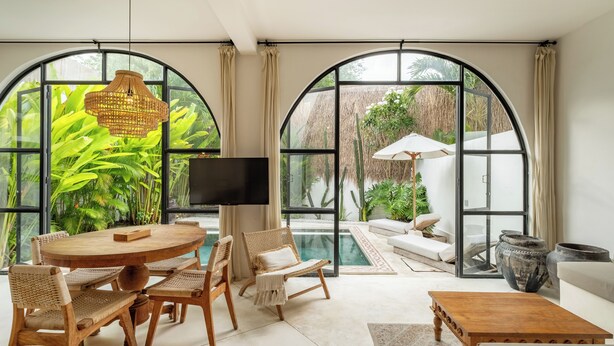 Amplia sala de estar con una mesa redonda de madera, sillas de mimbre, un sofá y una mesa de centro. Grandes ventanales de arco ofrecen vistas a exuberante follaje y una piscina.