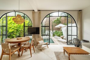 A spacious living area with a round wooden table, wicker chairs, a sofa, and a coffee table. Large arched windows offer views of lush greenery and a pool. -  Villa Akhila Canggu (Canggu)