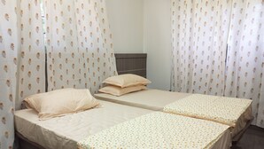 A room with two single beds, each with a beige mattress and matching pillows. The room has white curtains with a floral pattern and a wooden headboard.