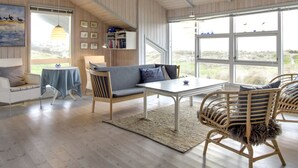 Ein geräumiges Zimmer mit Holzbalkendecke, einem großen Fenster, einer Couch, einem Tisch und Korbstühlen.