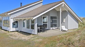 Ein Küstenhaus mit geneigtem Dach, großen Fenstern und einer Terrasse mit Gartenmöbeln.