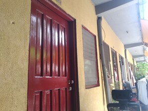 Une porte rouge sur un mur à texture, une fenêtre avec un cadre rouge et une chaise noire à l’extérieur.