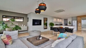 A modern living room with a sofa, coffee table, and a dining area with chairs and a kitchen in the background. - Amara 2 -designer Penthouse With Spectacular Views (Cape Town)