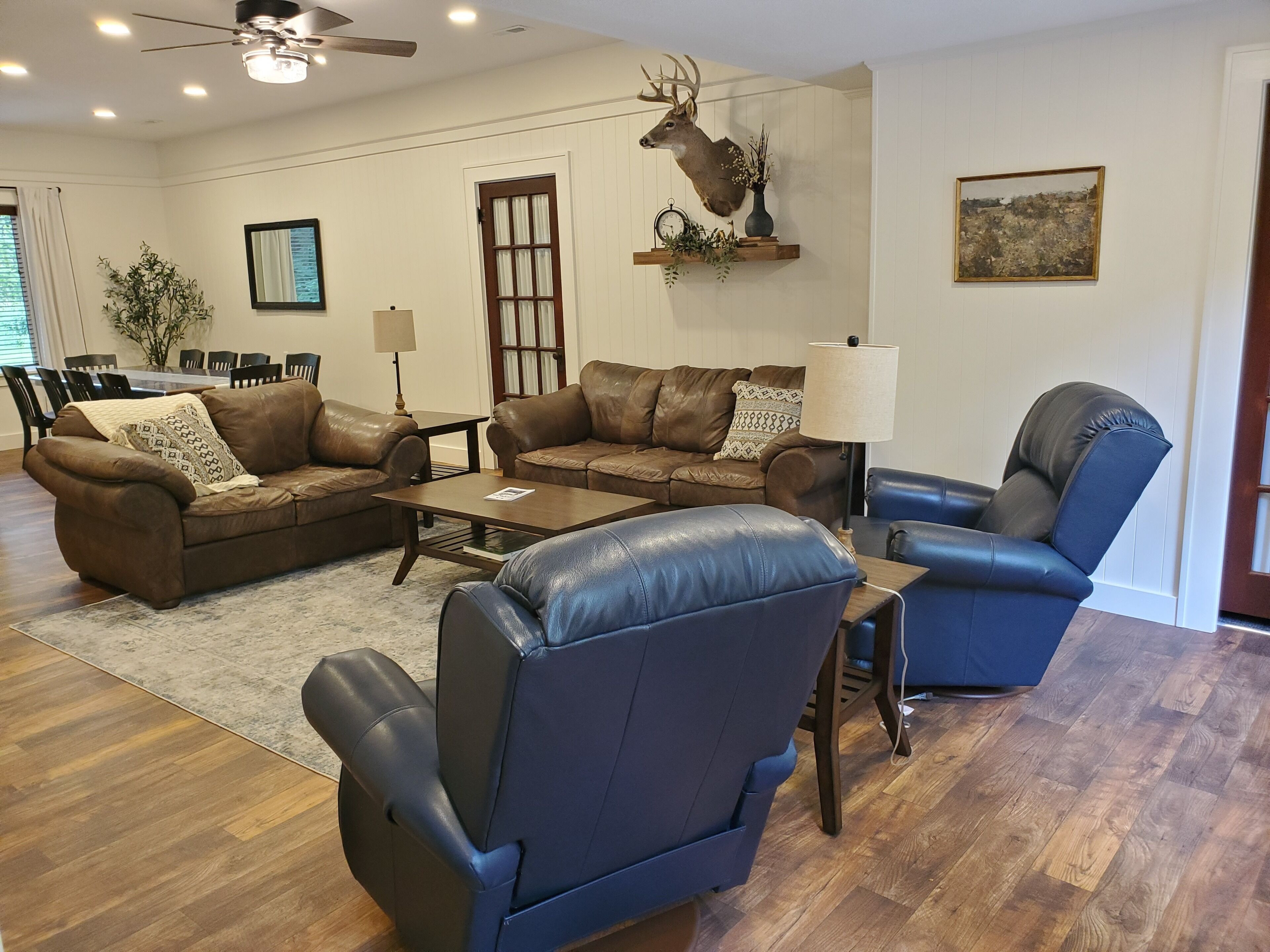 Un vaste salon agrémenté de deux canapés en cuir marron, d’un fauteuil en cuir bleu et d’une table basse en bois. Une tête de cerf décorative est accrochée au mur, et on aperçoit un coin repas au fond de la pièce.