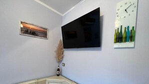 Una habitación con un televisor de pantalla plana en la pared, un cuadro enmarcado de un paisaje, una composición decorativa de botellas y un mantel a cuadros sobre una mesa auxiliar.