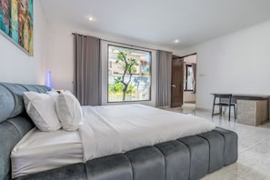 Une chambre moderne avec un grand lit, un bureau et une vue sur un immeuble et la verdure environnante.