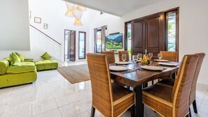 A dining area with a wooden table set for four, surrounded by wicker chairs. A green sofa is in the background, and a staircase leads to another room. - Villa Sky View - Seminyak (Seminyak)