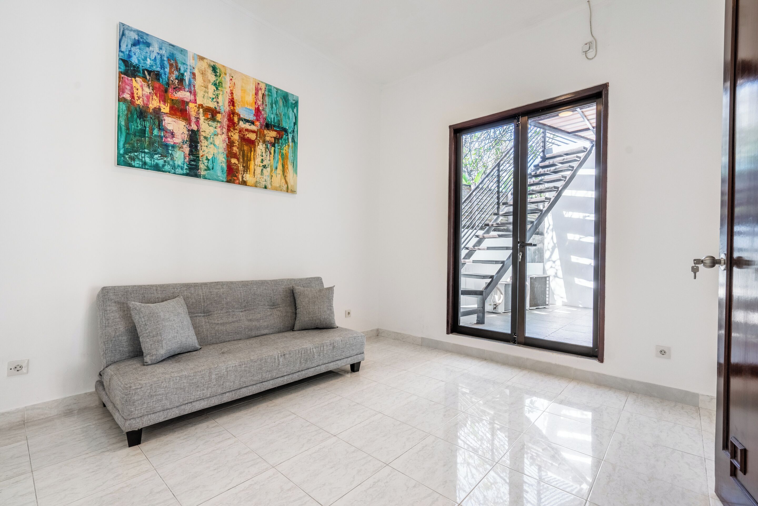 Una habitación amplia con un sofá gris, un cuadro abstracto colorido y un ventanal con vista a una escalera.