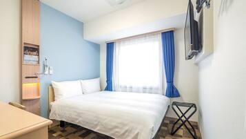 Quarto de hotel com cama, escrivaninha, cadeira, televisão na parede e janela com cortinas azuis.