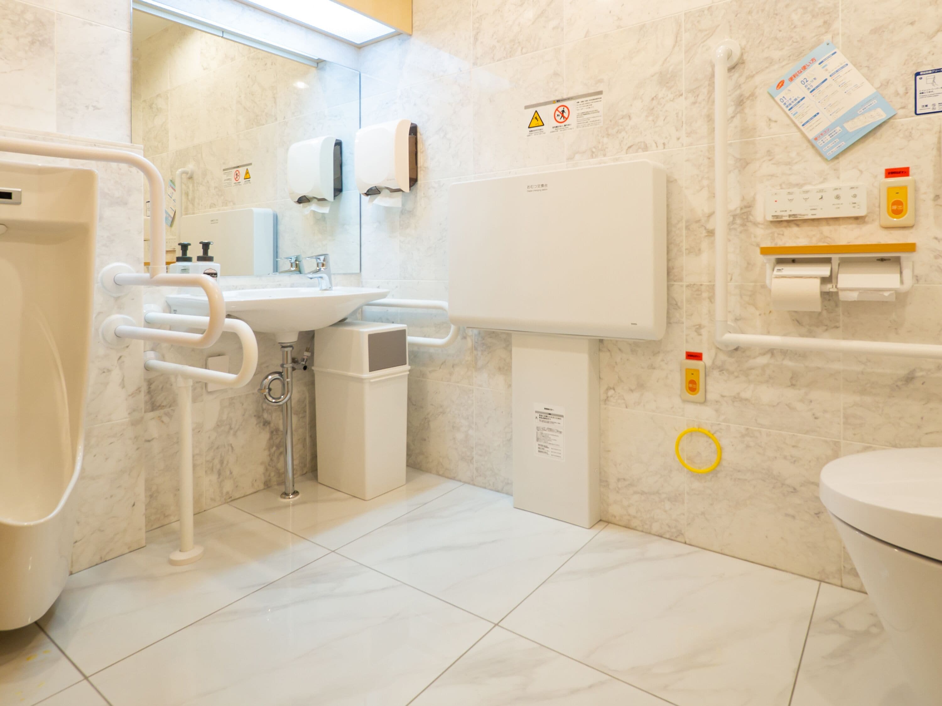 A modern bathroom with a sink, toilet, and bidet. There are hand dryers and a paper towel dispenser. The floor is tiled, and there are warning signs on the wall.