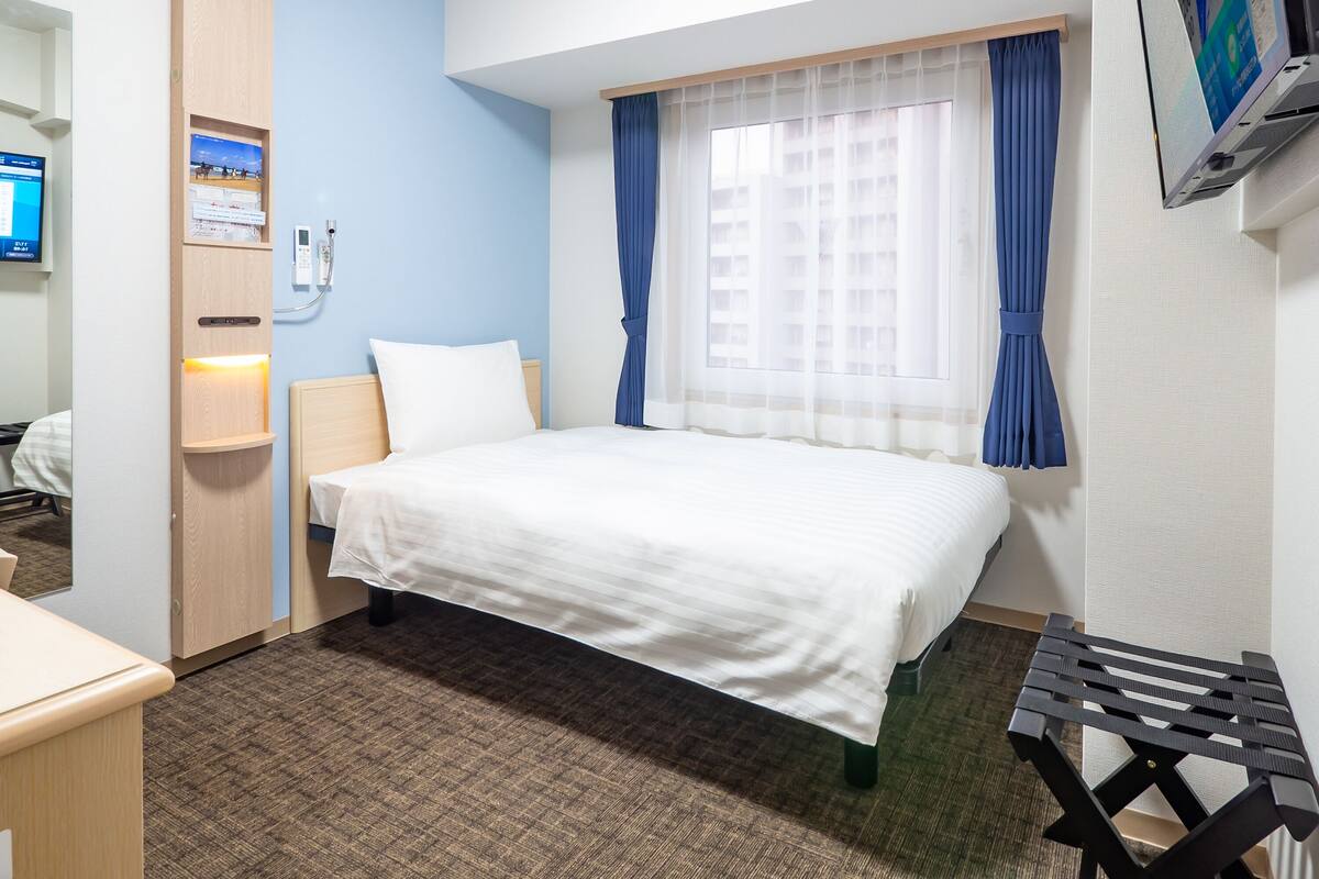 A hotel room with a bed, a desk, a chair, a TV, and a window with curtains.