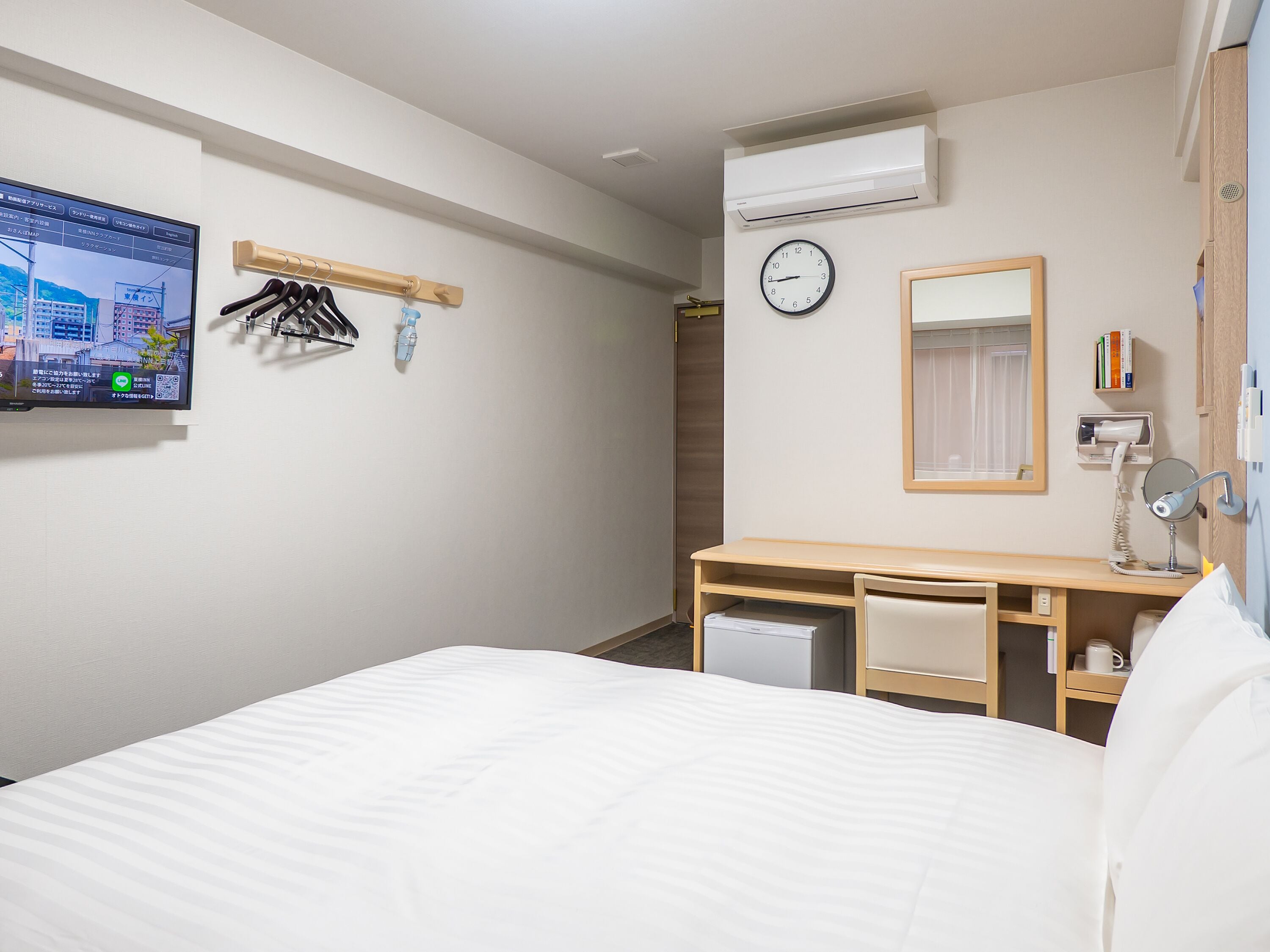 Une chambre d’hôtel avec un lit, un bureau, une chaise, une horloge, un miroir et un téléviseur fixé au mur.