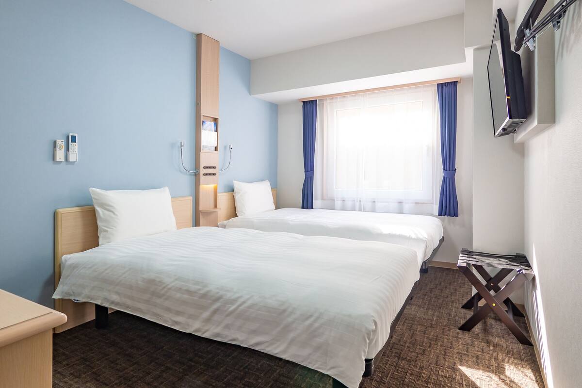 A hotel room with two beds, a wooden nightstand, a desk, a mirror, a window with curtains, and a flat-screen TV mounted on the wall.