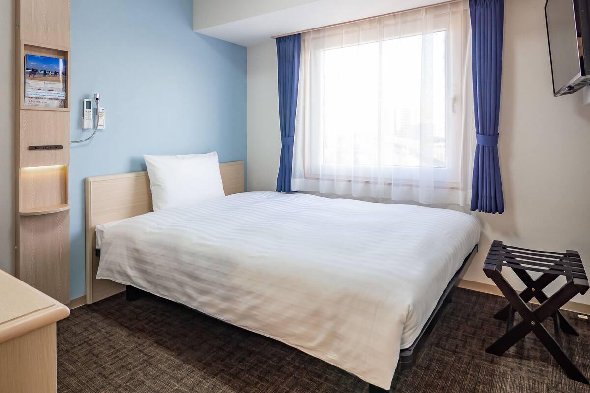 A hotel room with a bed, a desk, a small table, a TV, and a window with curtains.