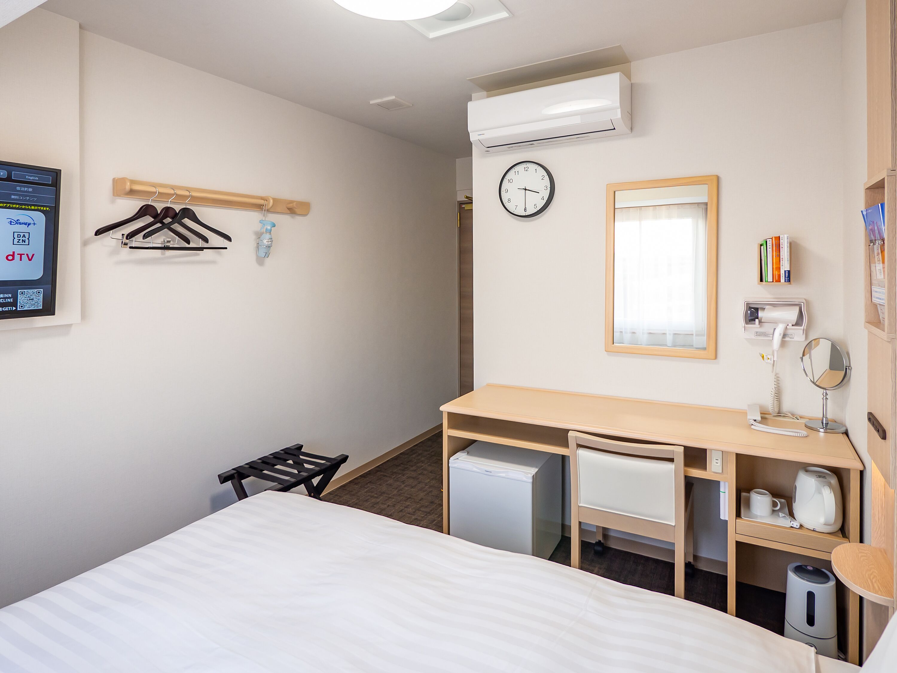 Habitación de hotel con cama, escritorio con espejo, refrigerador pequeño, calefacción, reloj y televisor montado en la pared.