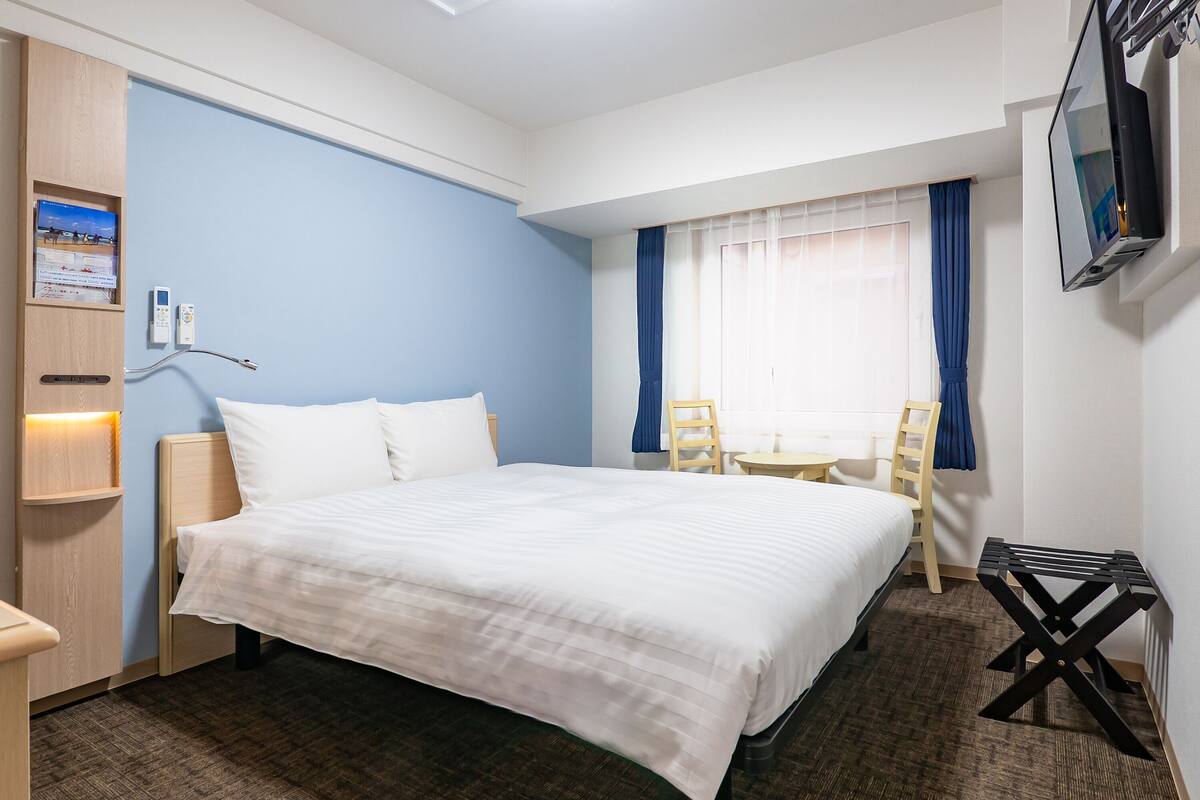 A hotel room with a bed, a desk, a chair, a TV, and a wall-mounted shelf.