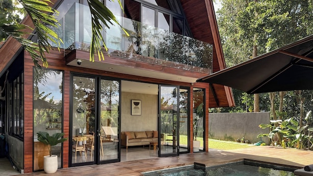 Une maison moderne dotée d’une piscine, d’une terrasse en bois et d’un salon entièrement vitré, comprenant un canapé et des fauteuils.