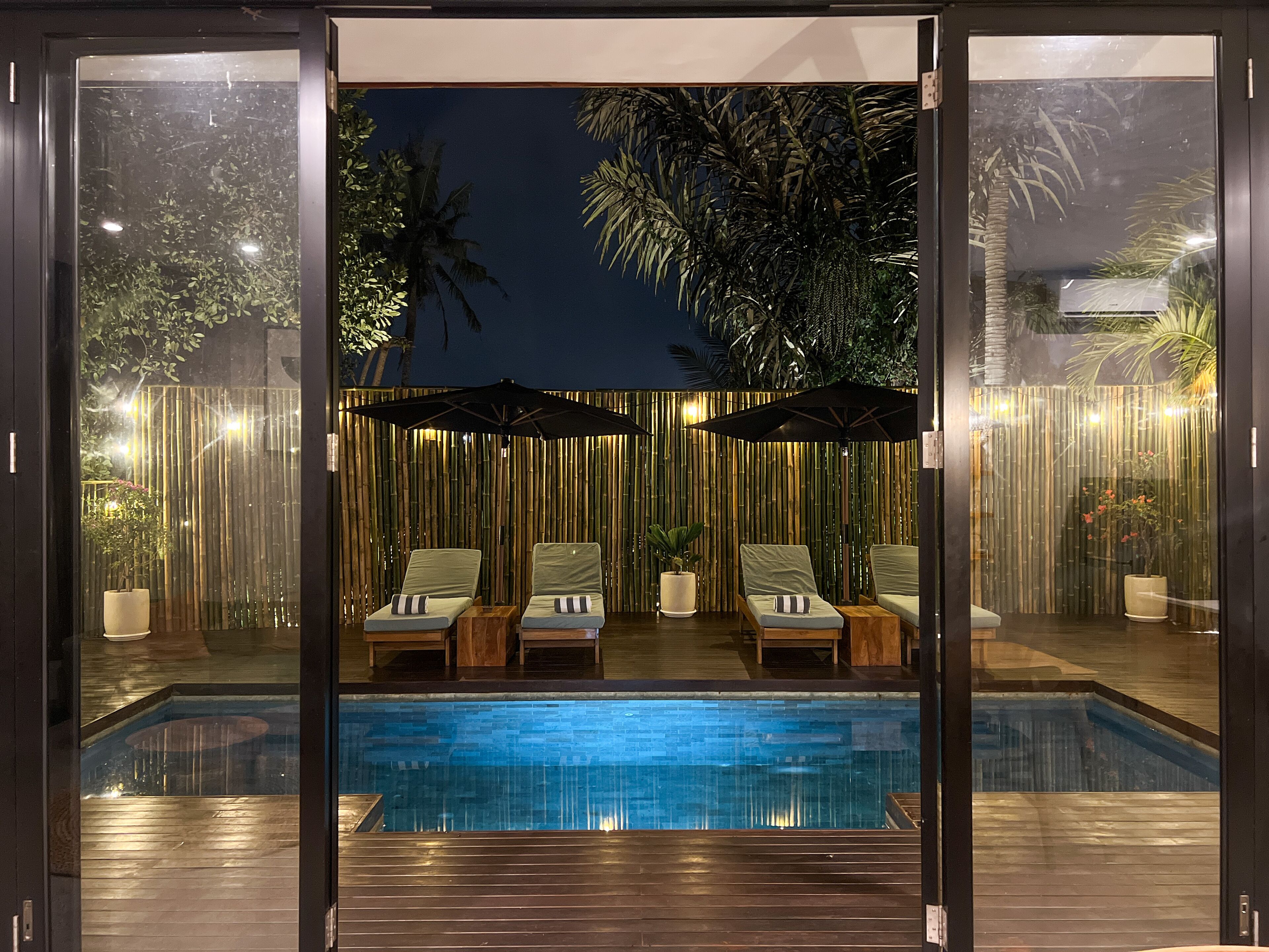 A poolside area with lounge chairs and umbrellas, surrounded by bamboo walls and palm trees.