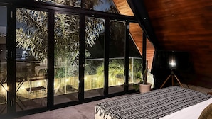 Une chambre avec une grande porte-fenêtre coulissante donnant sur un balcon, un lit avec une literie à motifs, une plante en pot et une lampe montée sur un trépied.