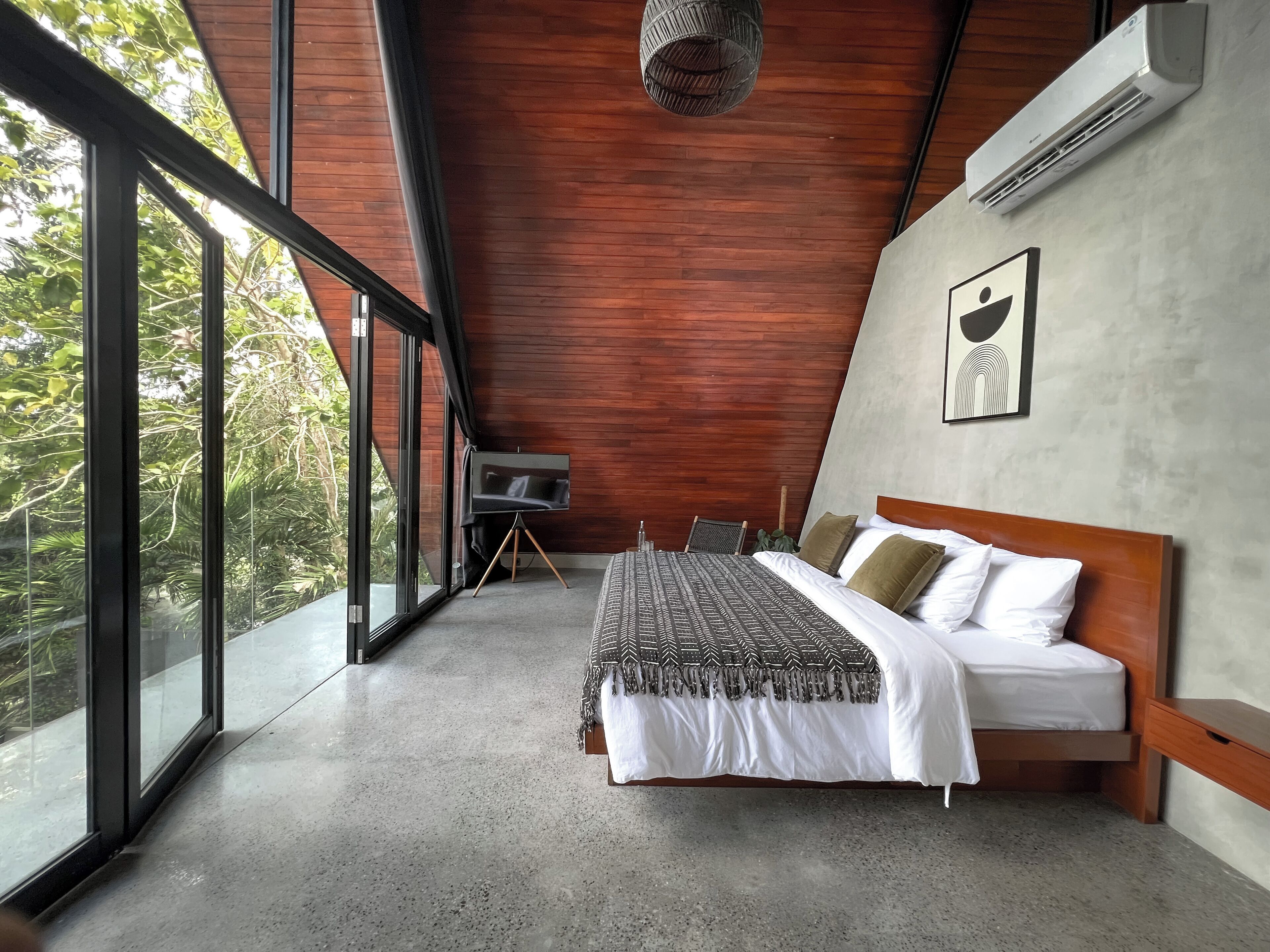 A bedroom with a large bed, a TV on a tripod, a chair, and a view of greenery through large windows.