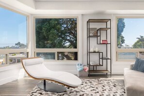 A modern living room with a white leather chair, a patterned rug, a bookshelf, and large windows offering a view of trees and buildings.