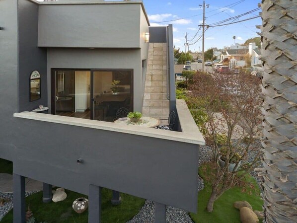 A modern two-story building with a flat roof, a balcony with a small table and chairs, and a stone wall.