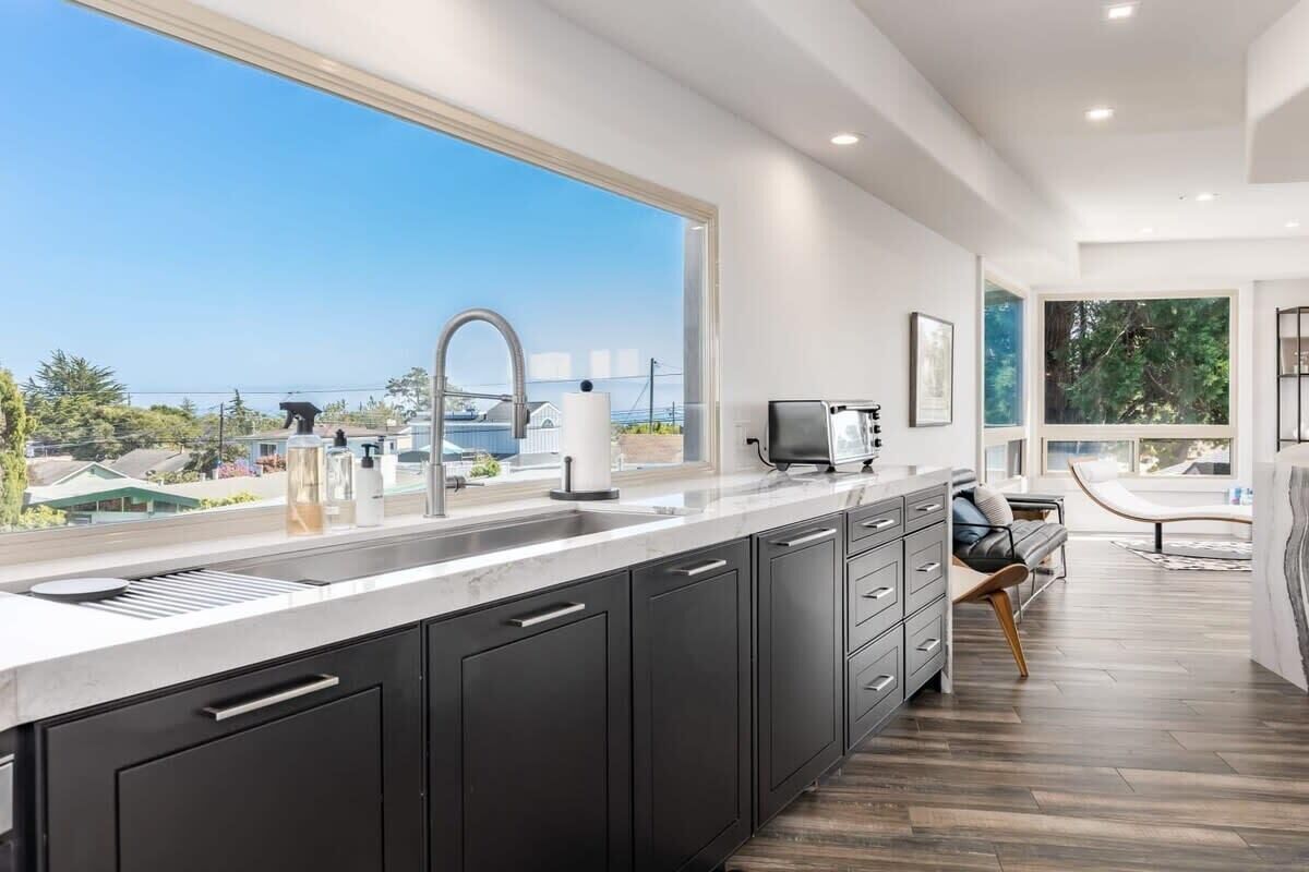 A modern kitchen with a large window, dark cabinetry, and a view of the sea.