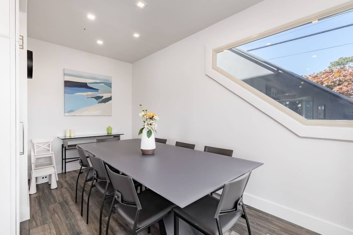 A modern dining area with a long table, chairs, a vase with flowers, a painting on the wall, and a skylight.