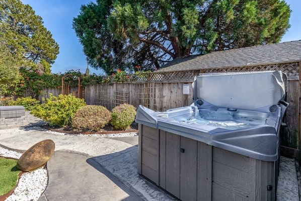 A hot tub in a backyard with a wooden fence, a large tree, and a stone pathway.