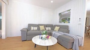 A modern living room with a grey sectional sofa, a round coffee table, and a window with plants. - Melbourne 4BRM Staycation Parkside (Hallam)