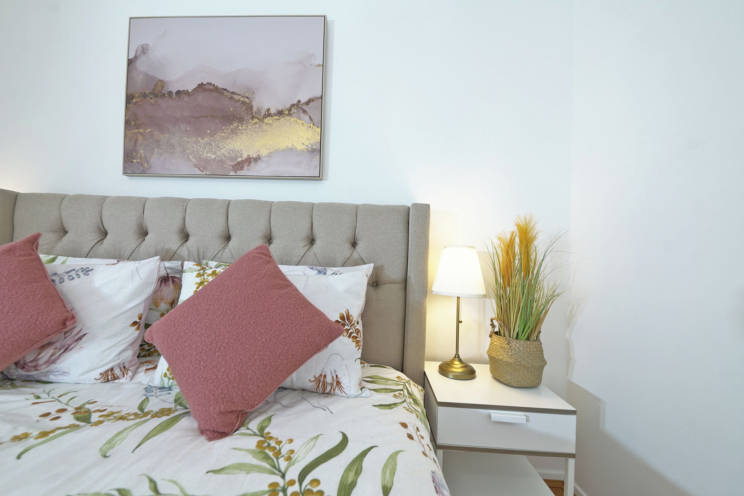 A neatly made bed with floral bedding and two pink pillows, a bedside table with a lamp and a plant, and a wall-mounted abstract painting.