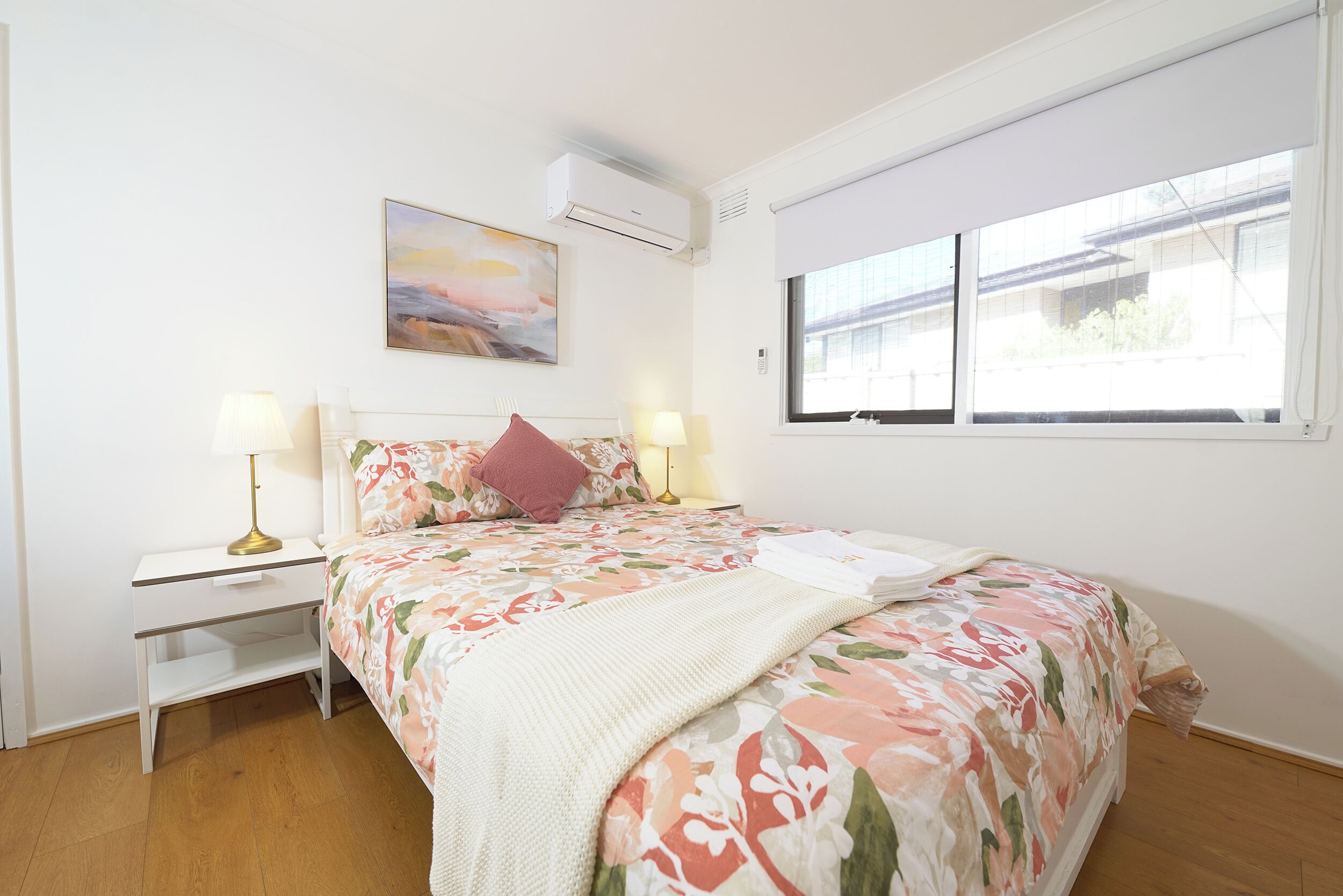 Un dormitorio con una colcha estampada, una mesita de noche con lámpara, una ventana con persianas y un equipo de aire acondicionado en la pared.