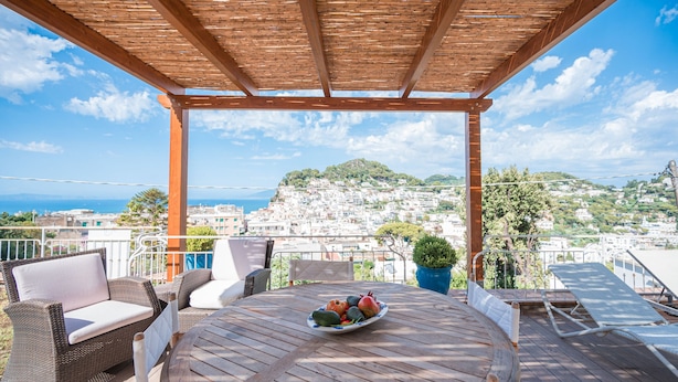 A wooden deck with wicker furniture and a table with fruit. A view of a hillside town is visible through the pergola. - Villa Albachiara in Capri (Capri)