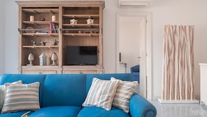 A living room with a blue sofa, a wooden cabinet with shelves, a television, and a tall, striped curtain. - Villa Albachiara in Capri (Capri)