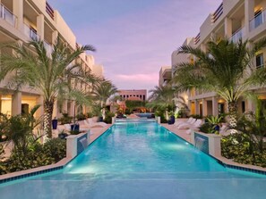 A swimming pool with lounge chairs and palm trees, surrounded by hotel buildings.