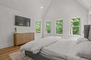 Une chambre à coucher comprenant un lit, une commode en bois, un téléviseur à écran plat et plusieurs fenêtres donnant sur un paysage verdoyant.