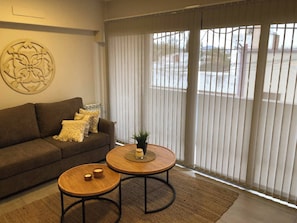 A living room with a brown sofa, two round wooden tables, a decorative wall piece, and a large window with vertical blinds. - Apartment First Flor Excellent Location (Salta)