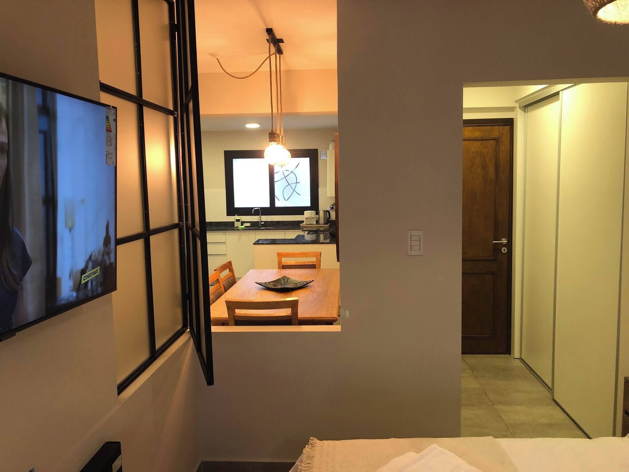 A room with a television mounted on the wall, a dining area with a table and chairs, and a kitchen visible through an open door.
