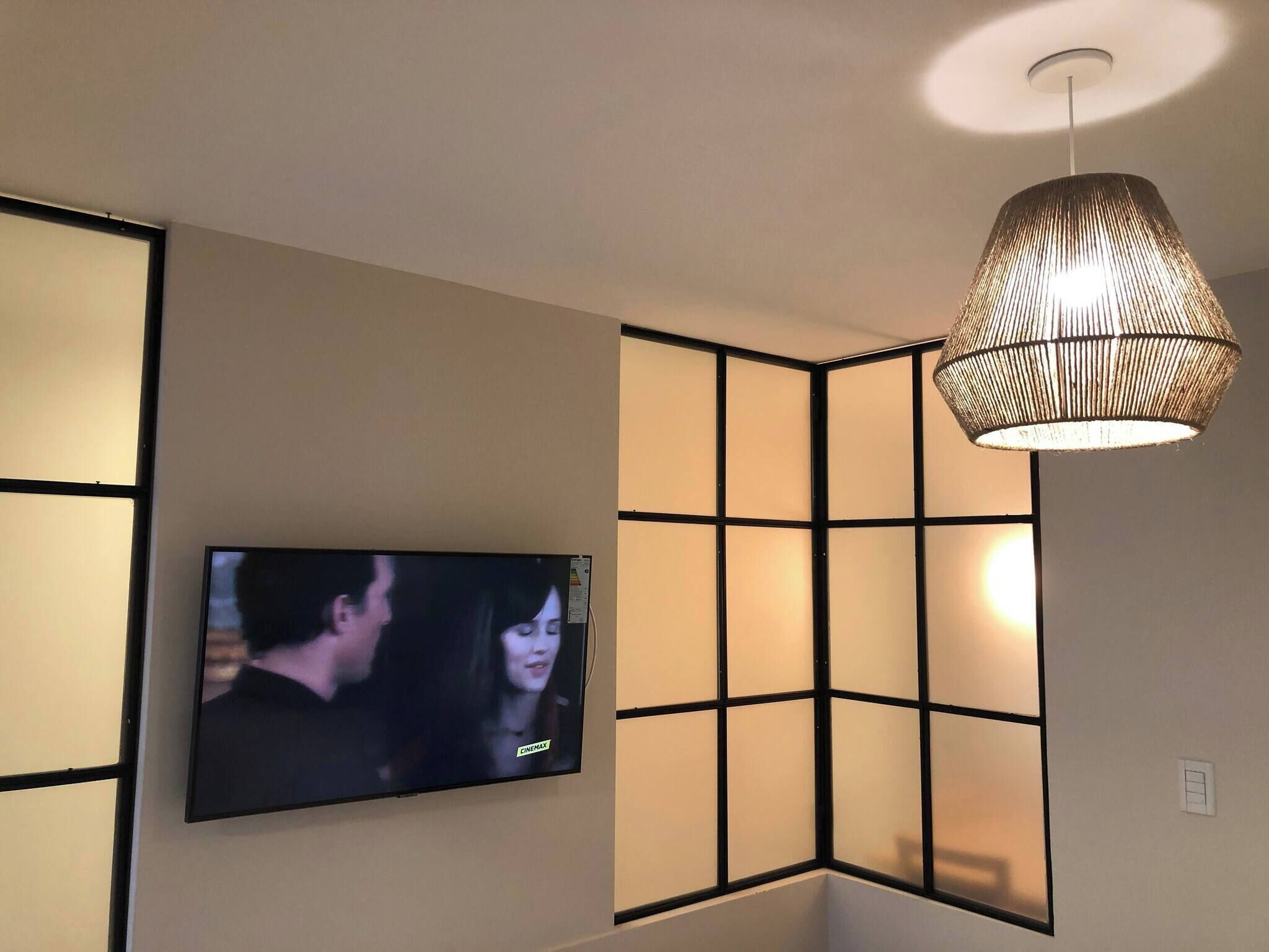 A room with a flat-screen TV mounted on the wall, a woven pendant light, and a partition with a grid pattern.