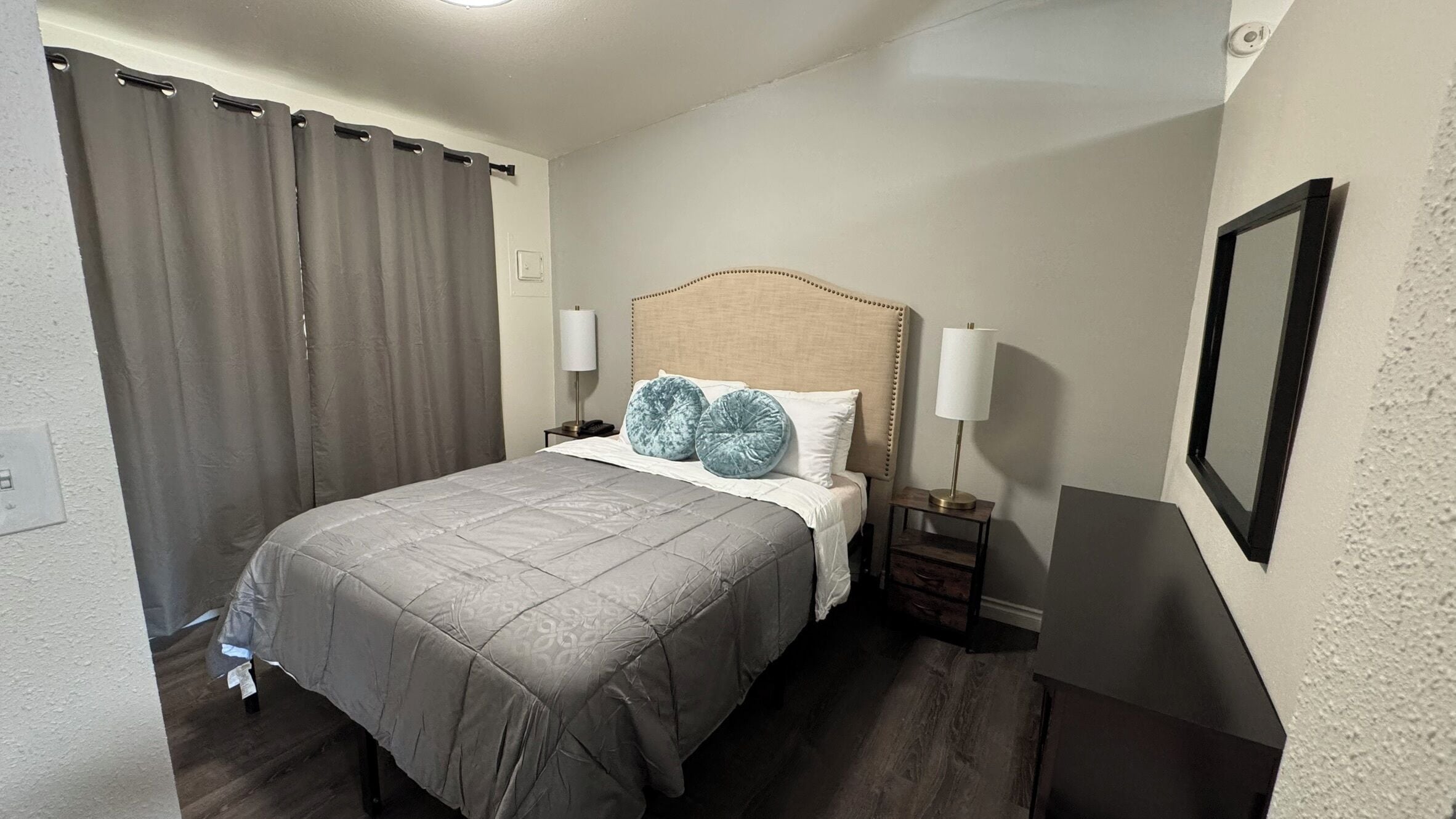 Habitación de hotel con cama, cortinas, mesita de noche, dos lámparas y un cuadro en la pared.