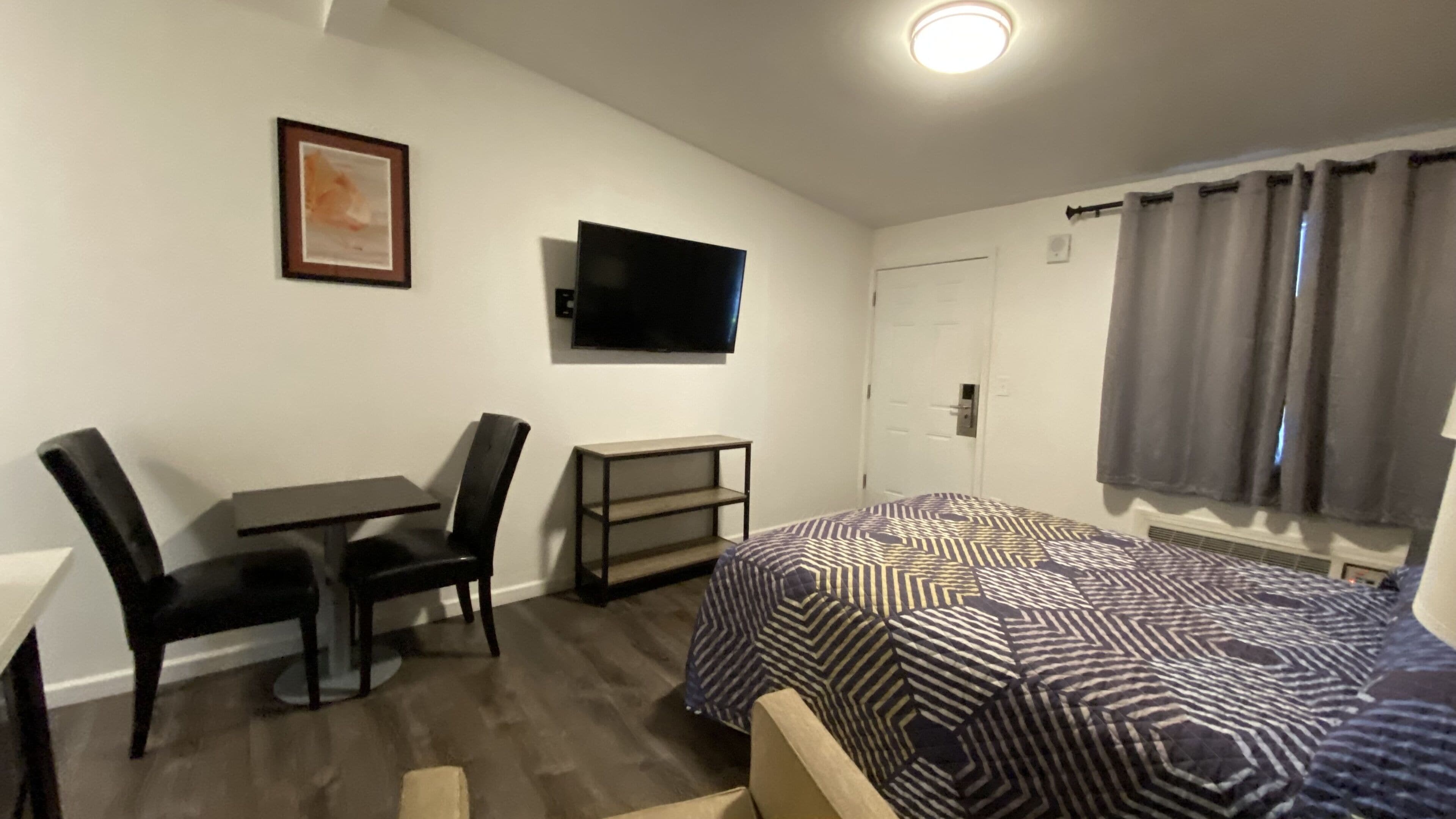 Una habitación pequeña y luminosa con una cama, un escritorio, una silla, un televisor y una ventana con cortinas.
