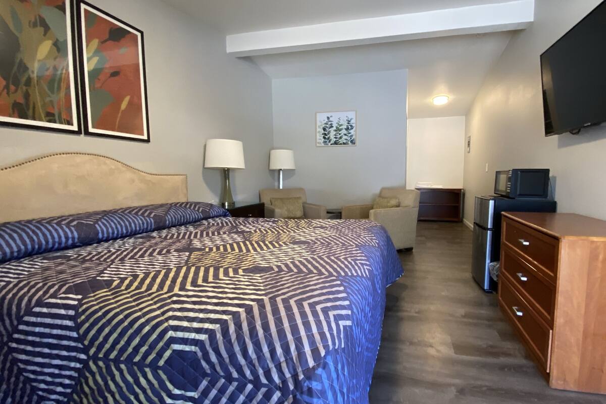 A hotel room with a large bed, two chairs, a wooden dresser, a television, and a refrigerator.