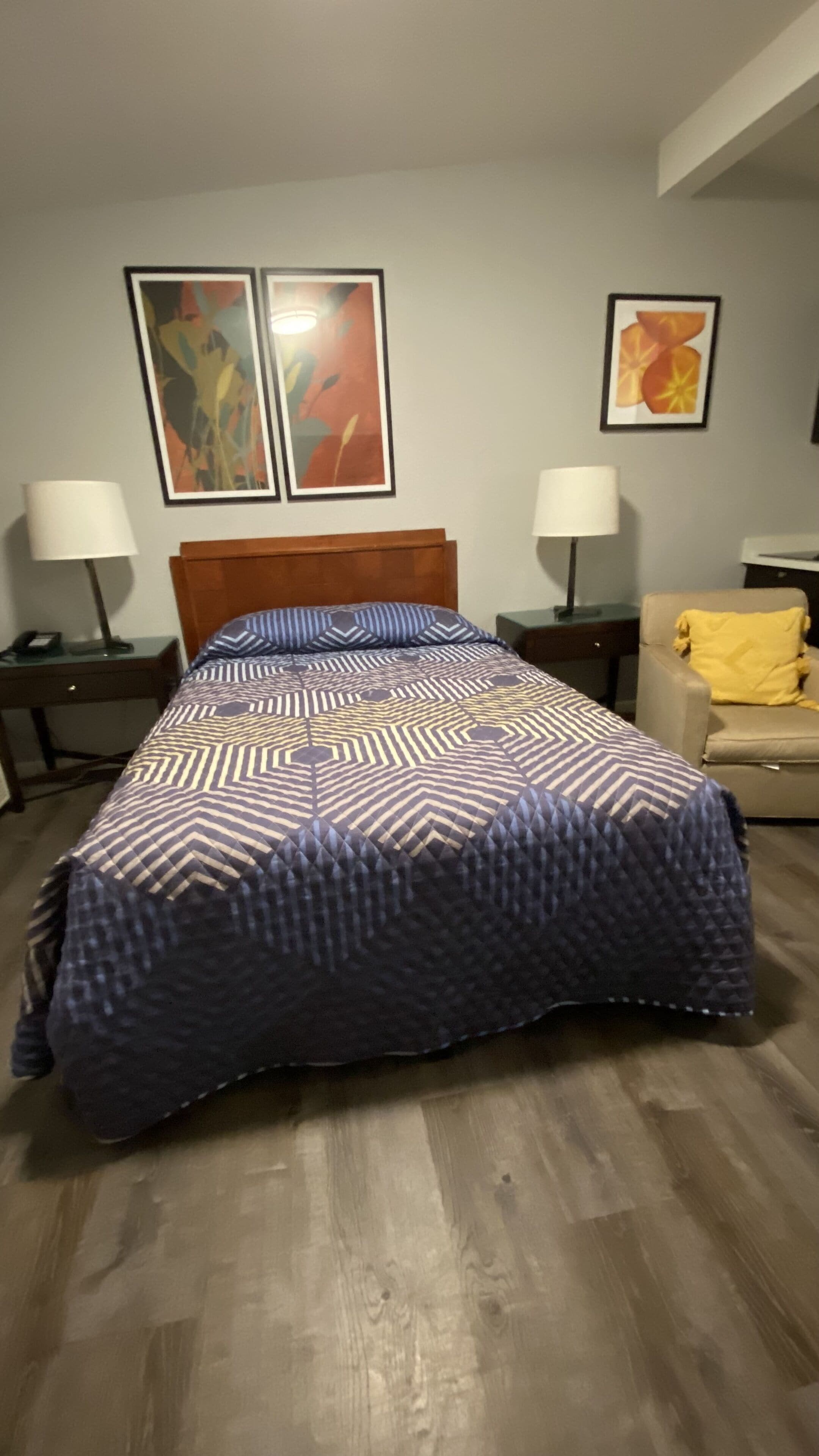 A neatly made bed with a patterned bedspread, two bedside tables with lamps, a chair with a yellow throw, and two abstract paintings on the wall.